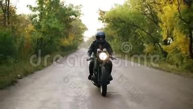秋天，一个戴着墨镜和皮夹克的年轻人骑着摩托车骑在柏油路上的正面景色